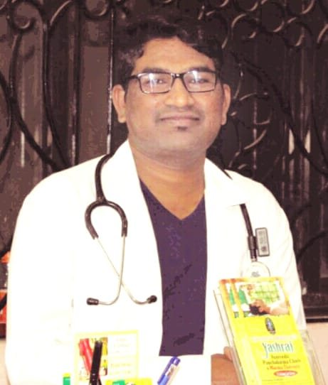 Dr. Raju Bhusanar, BAMS — in his white coat with stethoscope, holding an Ayurvedic Panchakarma text at the clinic